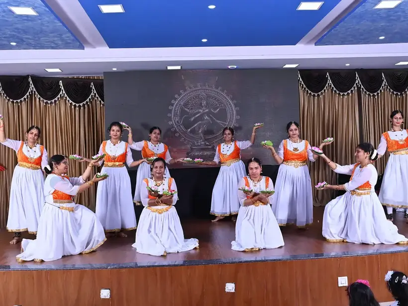 Students in white and orange costumes performing a classical dance on the auditorium stage