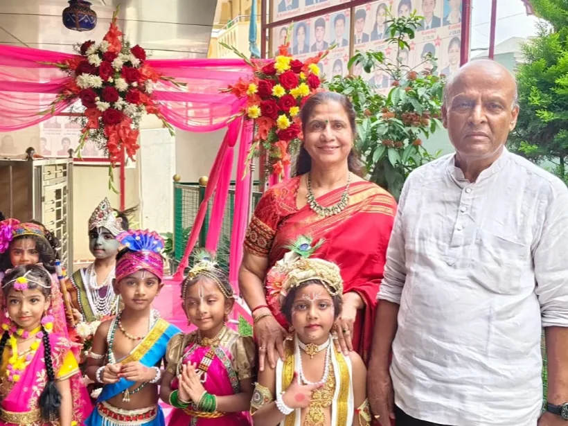 A group of children dressed as Krishna and Radha for Krishna Janmashtami, with two senior staff members, beneath a flower-decorated mandap