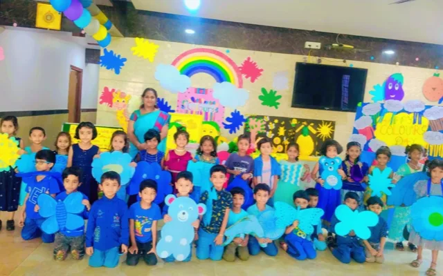 Kindergarten students dressed in blue holding blue paper craft cutouts — butterflies, flowers and a teddy bear — with a teacher for Colour’s Day