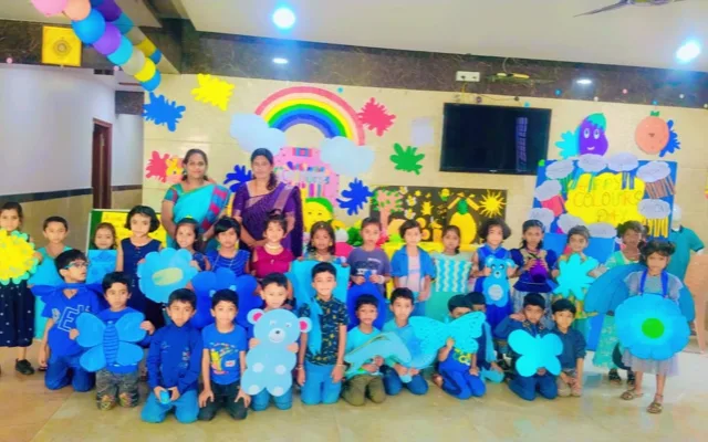 Students and two teachers dressed in blue posing in front of a "Happy Colours Day" backdrop with yellow and blue craft cutouts