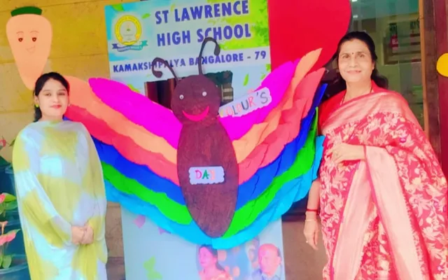 Two staff members beside a large rainbow-coloured butterfly display and the St. Lawrence High School signage for Colour’s Day