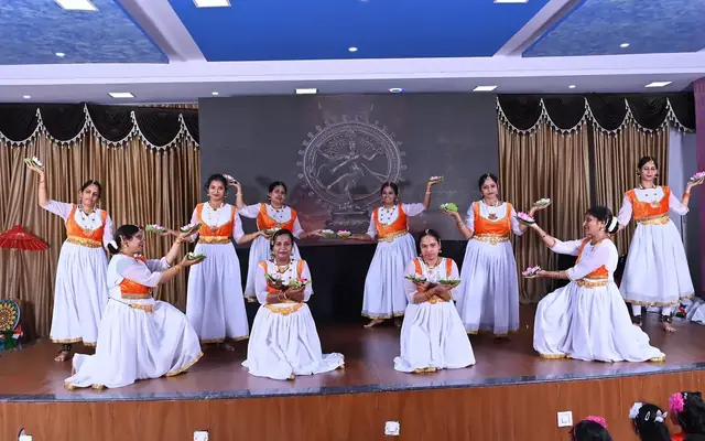 Students in white and orange costumes performing a classical dance on the auditorium stage
