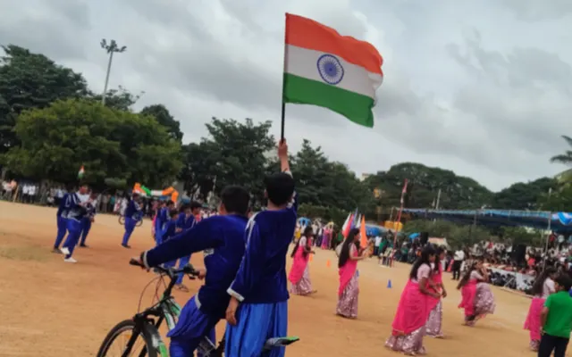 A student cycling while holding the national flag aloft as classmates perform a display behind